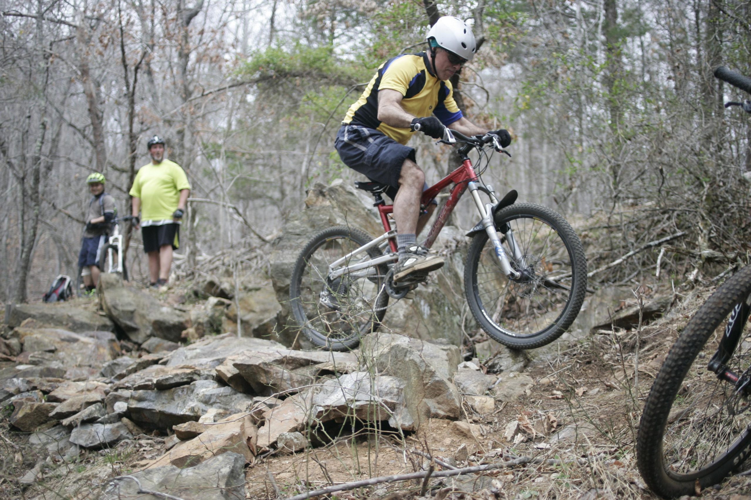 A mountain biker in a yellow and blue jersey and white helmet is jumping over a rocky terrain, while two other cyclists observe from a higher vantage point in a wooded area. The setting is surrounded by trees with sparse leaves, indicating an early spring or late fall season. Mountain Laurel Trails mountain bike trail.