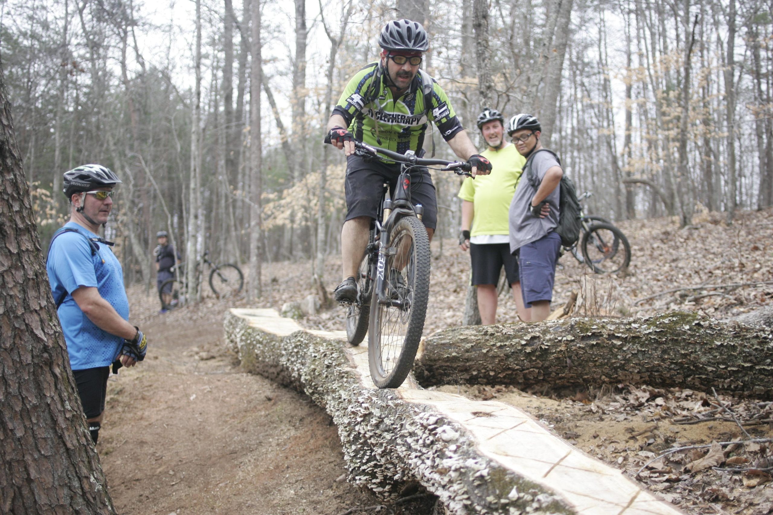 A cyclist balances on a log while riding a mountain bike on a nature trail, with three onlookers watching. The scene is set in a wooded area with leafless trees and a dirt path, suggesting an outdoor biking event. Mountain Laurel Trails mountain bike trail.