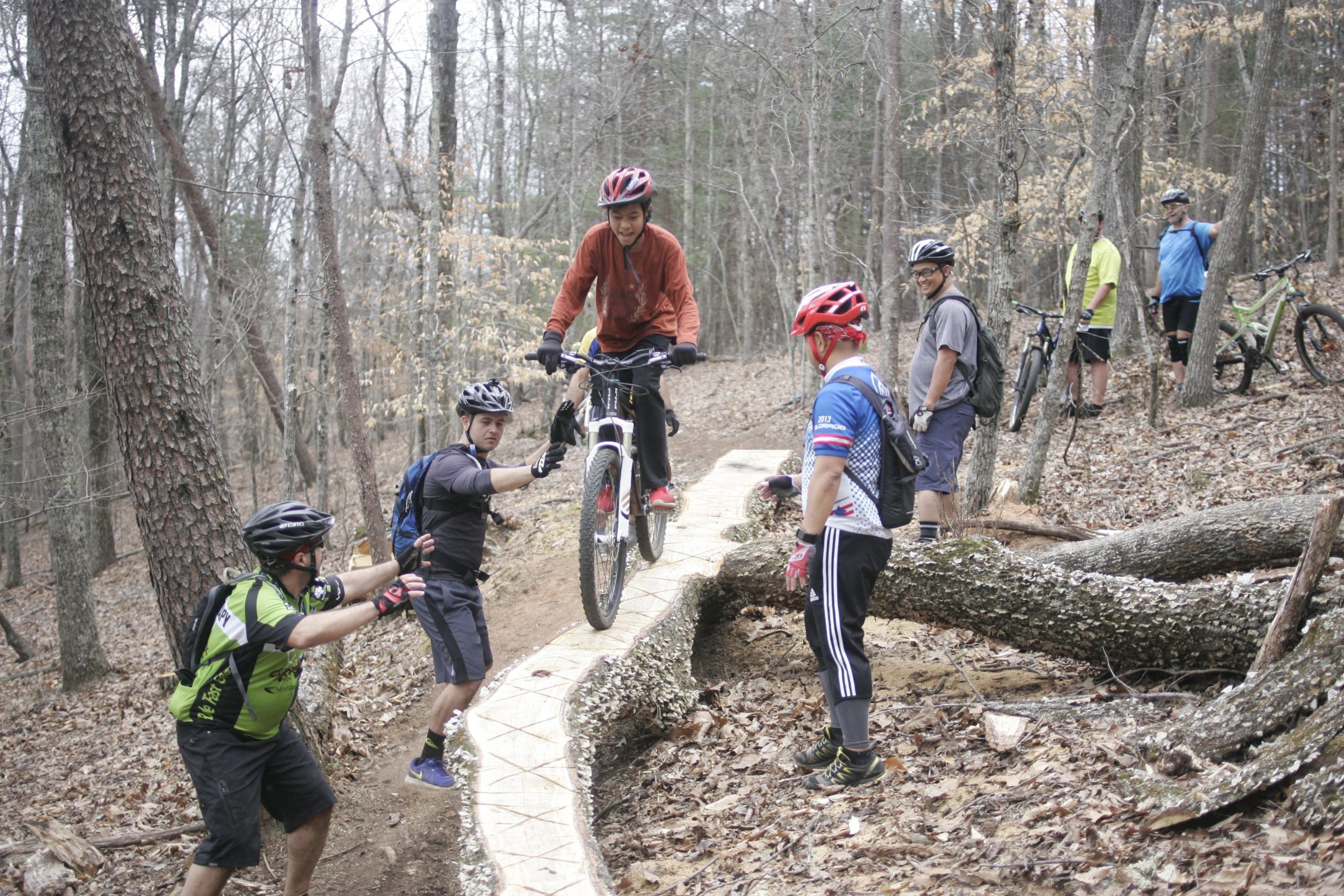 A group of mountain bikers gathered on a wooded trail, with one cyclist balancing on a narrow wooden path while others assist and observe. The scene captures a moment of camaraderie and teamwork amid the natural surroundings of trees and fallen leaves. Mountain Laurel Trails mountain bike trail.