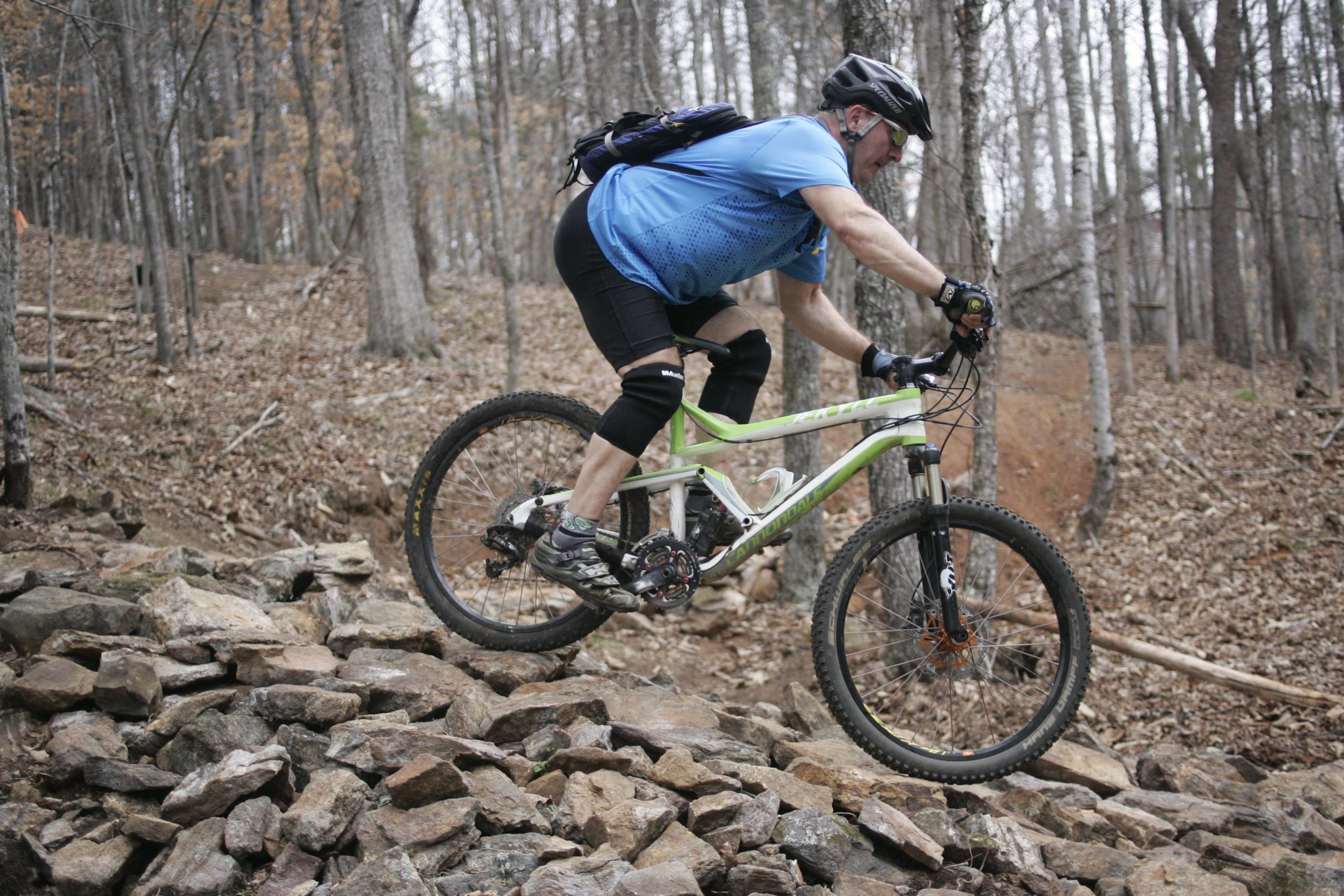 A mountain biker navigating over a rocky terrain in a forested area. The rider, wearing a helmet and protective gear, is in a focused position as they maneuver their bicycle across the uneven surface of stones, with trees and fallen leaves in the background. Mountain Laurel Trails mountain bike trail.
