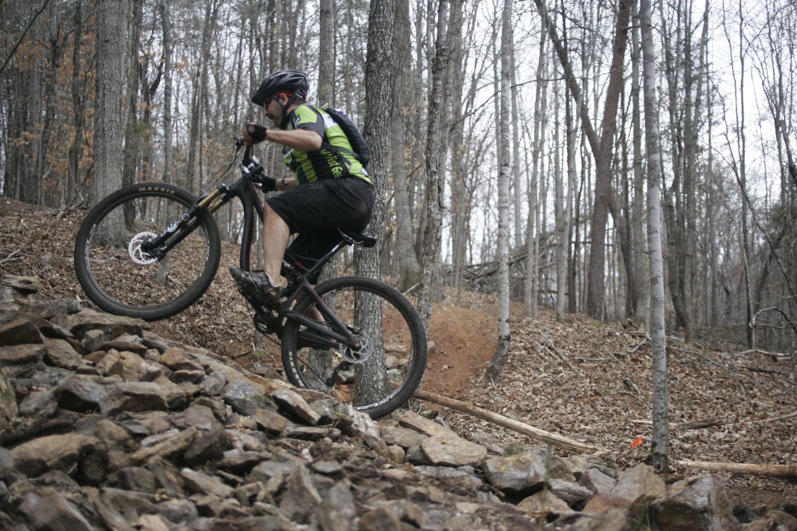 A mountain biker navigating over rocky terrain in a wooded area, with trees in the background and fallen leaves on the ground. The biker is wearing a black helmet and a bright green jersey, lifting the front wheel of the bike as he climbs the rocks. Mountain Laurel Trails mountain bike trail.