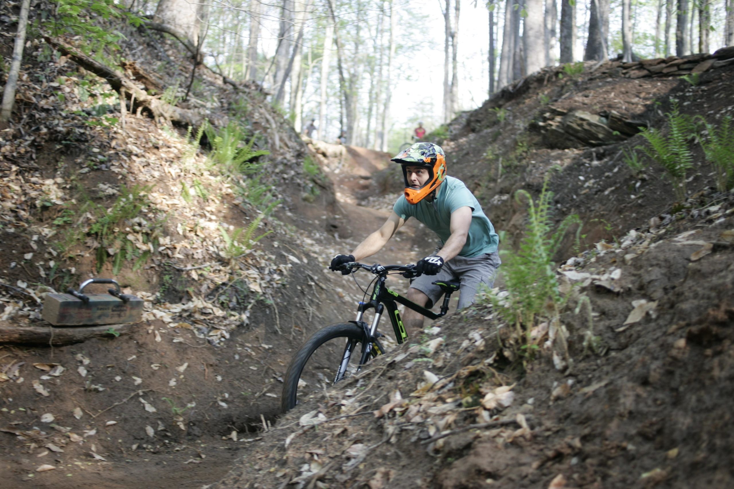 A mountain biker riding down a dirt trail surrounded by trees and ferns. The rider is wearing an orange helmet and gloves, and is focused on navigating the path. In the background, another biker can be seen on the trail. Salem Lake mountain bike trail.