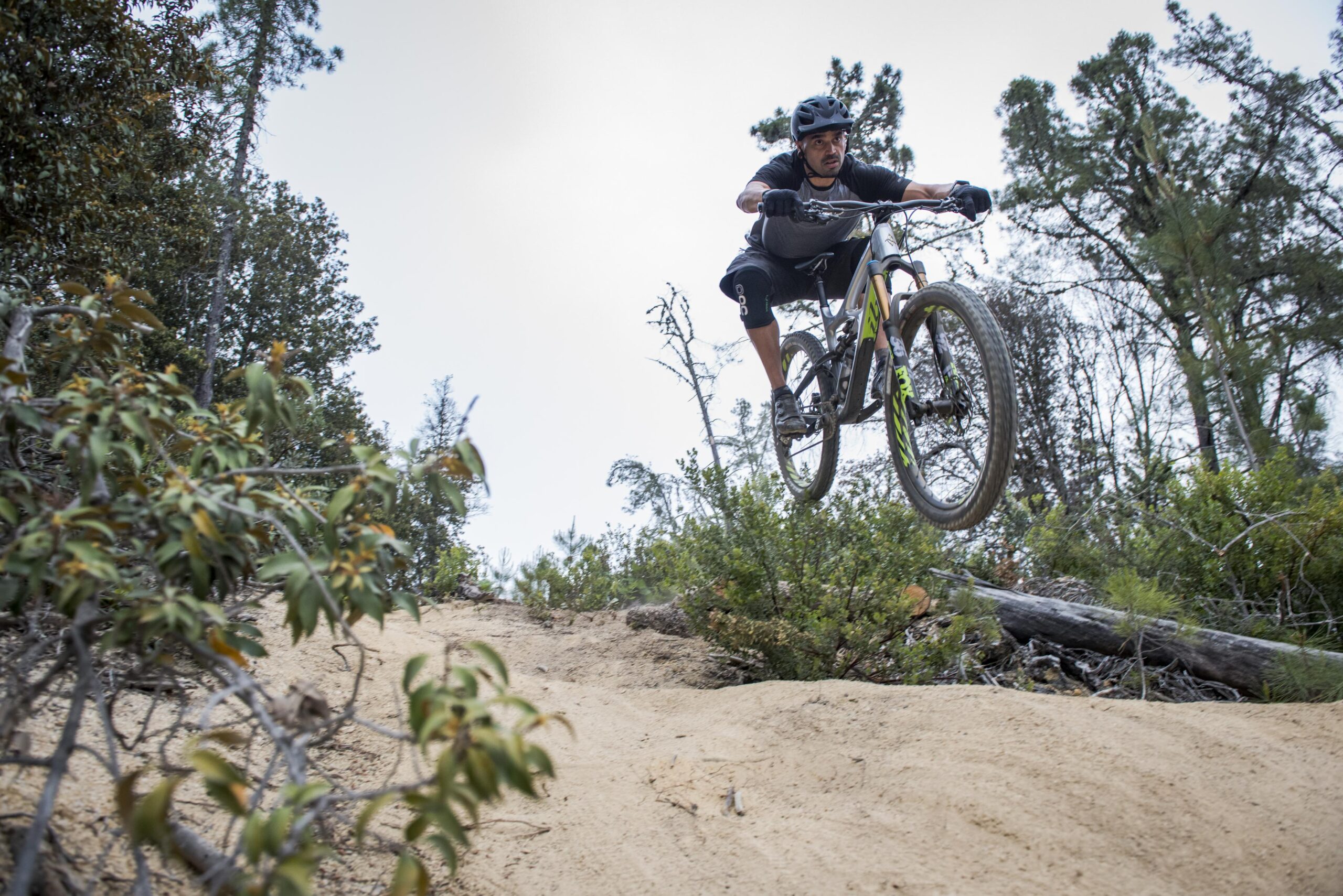 Ibis Mojo HD4: A mountain biker in mid-air performing a jump over a sandy trail surrounded by greenery and trees. The rider is wearing a helmet and sporty attire, showcasing a dynamic pose as they navigate the terrain.