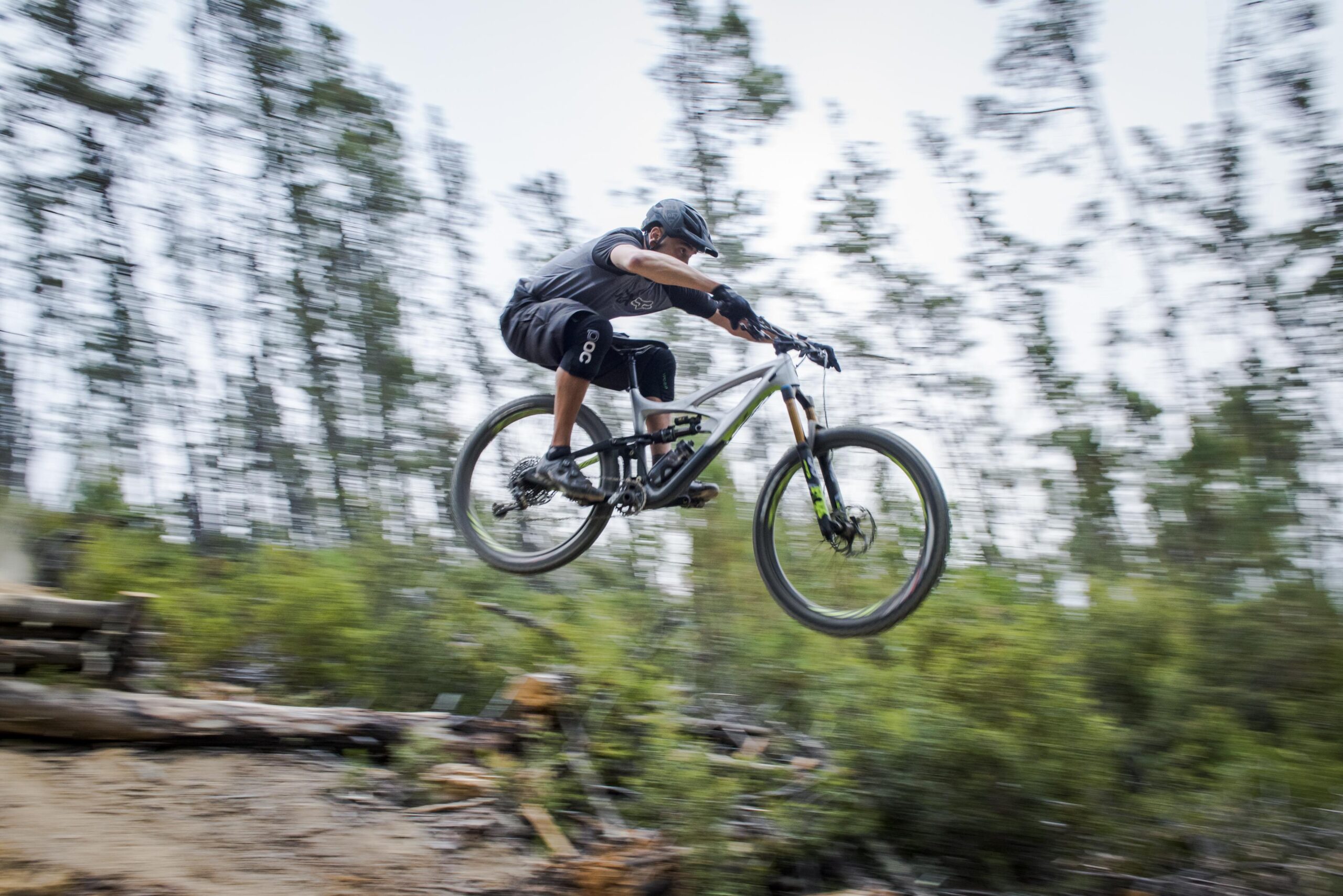 Ibis Mojo HD4: A person riding a mountain bike leaps off a dirt ramp, surrounded by tall trees and greenery. The image captures the motion and excitement of mountain biking, with a blurred background emphasizing speed. The rider is dressed in athletic gear and wearing a helmet.