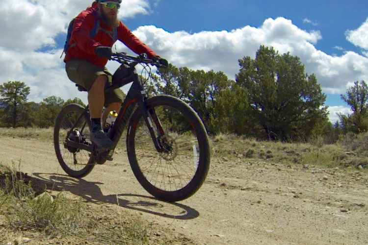 A person riding a mountain bike on a dirt trail, wearing a red jacket, helmet, and sunglasses. The scene features a backdrop of trees and a partly cloudy sky.