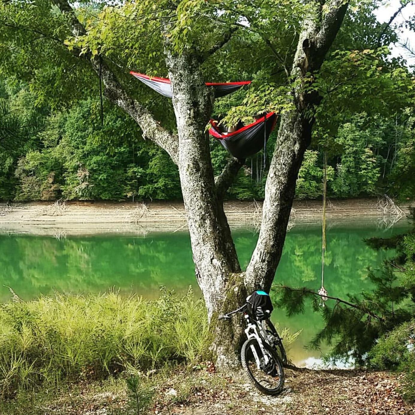 A serene lakeside scene featuring two hammocks suspended from a tree, with a mountain bike leaning against the tree trunk. The calm, green waters of the lake reflect the greenery surrounding it, and a rope hangs from a branch nearby. Brush Creek mountain bike trail.