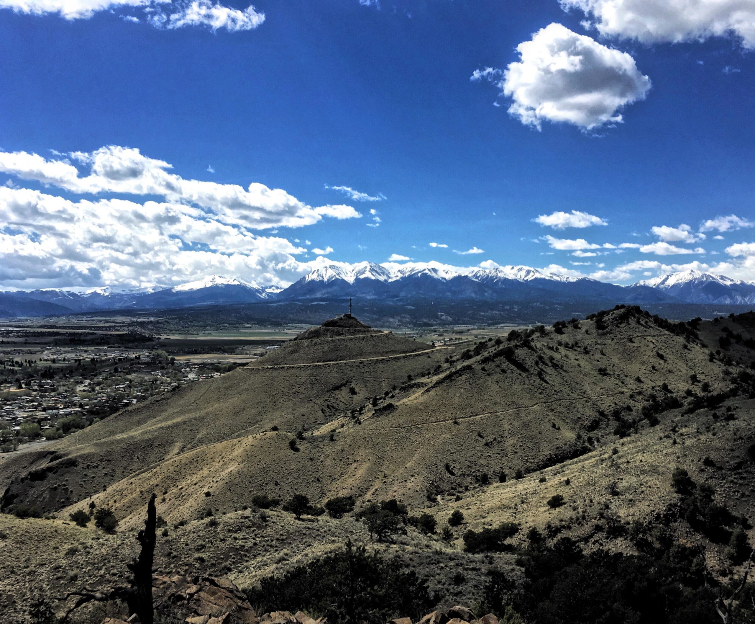 A panoramic view of rolling hills with sparse vegetation, leading to a distant mountain range capped with snow. The sky is bright blue with scattered clouds. In the foreground, a hill features a small cross at its peak, and a town is visible in the valley below, surrounded by green fields. Arkansas Hills mountain bike trail.