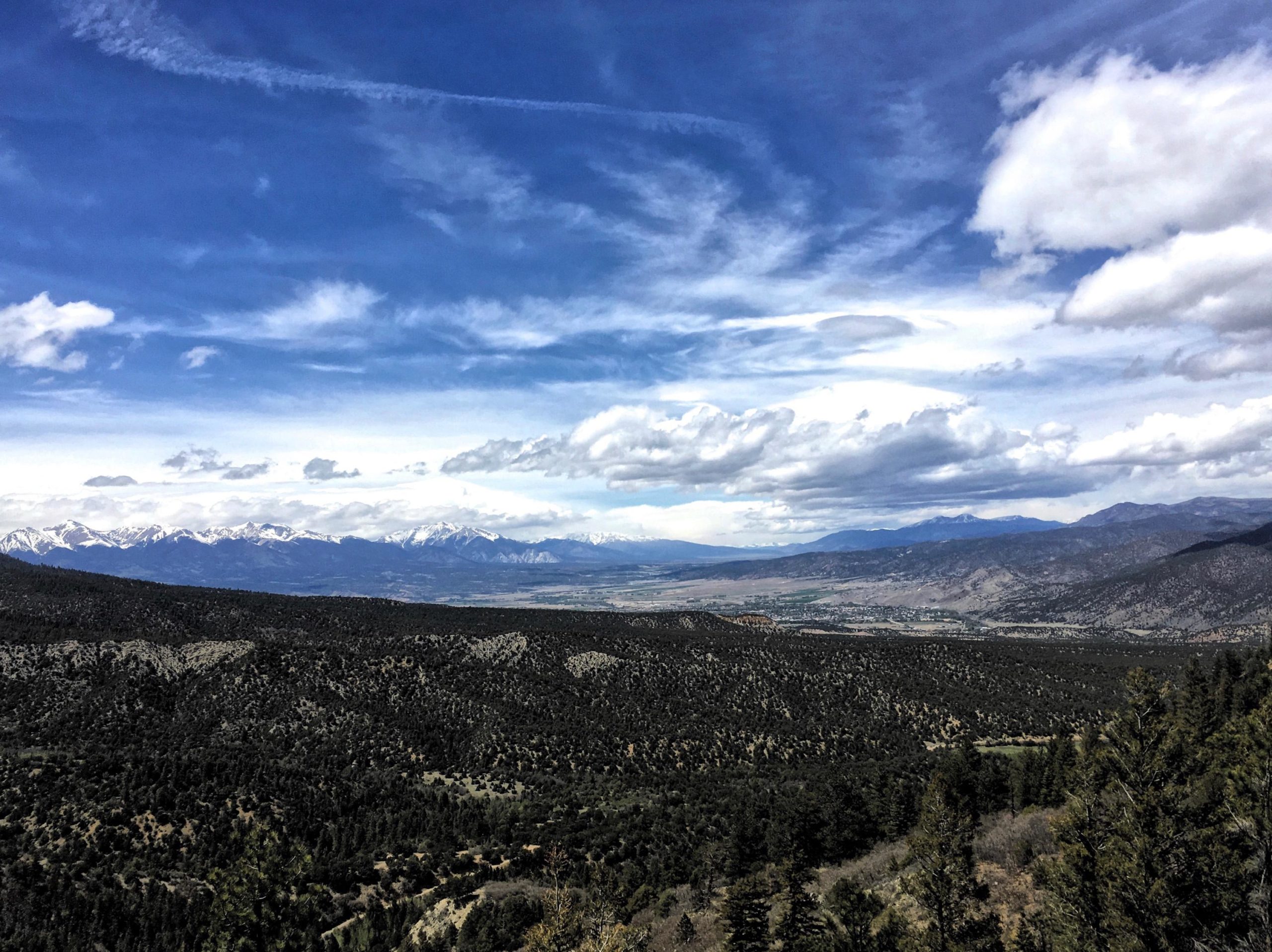 A panoramic view of a mountainous landscape under a bright blue sky, featuring scattered clouds. Snow-capped peaks rise majestically in the distance, while the foreground displays rolling hills covered with green foliage and a mix of trees. The valley below is dotted with patches of light and shadow, indicating a vibrant natural environment. Road #101.A mountain bike trail.