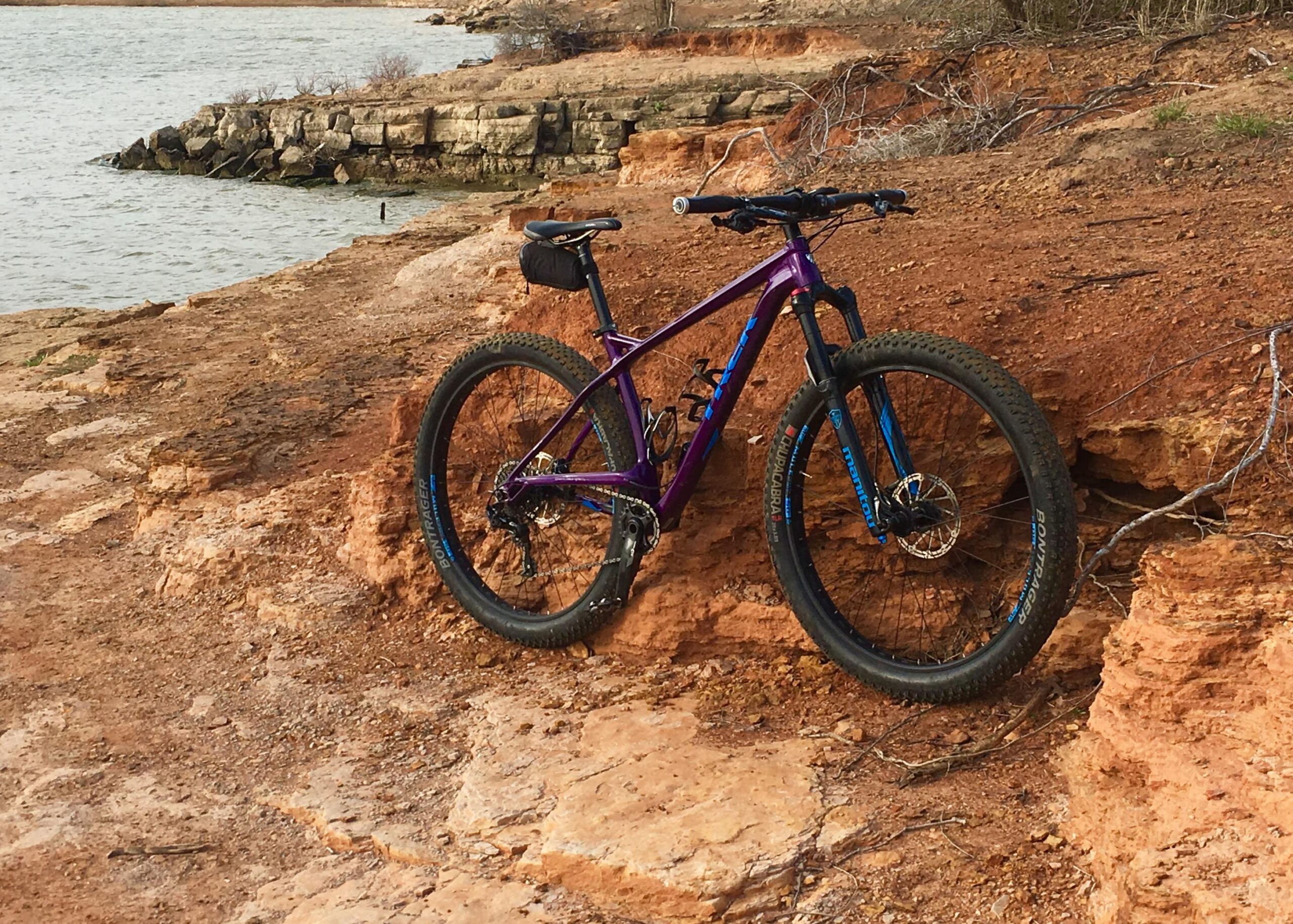 Trek Stache: A purple mountain bike resting on the rocky shore near a lake, with a background of water and a partially wooded landscape. The ground is uneven, showcasing reddish-brown earth and scattered twigs.