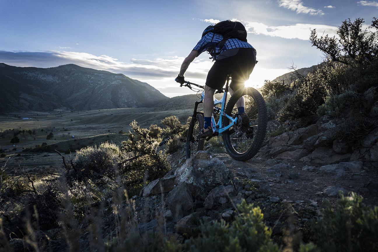 Terrene McFly: A mountain biker navigating a rocky trail during sunset, with mountains and a valley in the background. The cyclist is wearing a blue plaid shirt and riding a blue bike, showcasing the beauty of outdoor adventure.