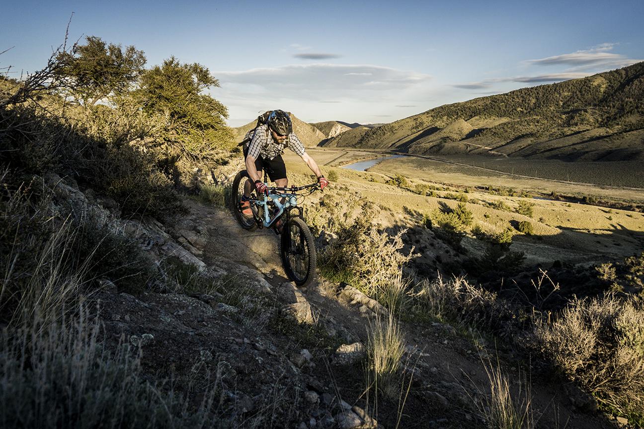 Terrene McFly: A mountain biker navigating a rocky trail in a mountainous landscape during sunset, with hills and a river visible in the background.