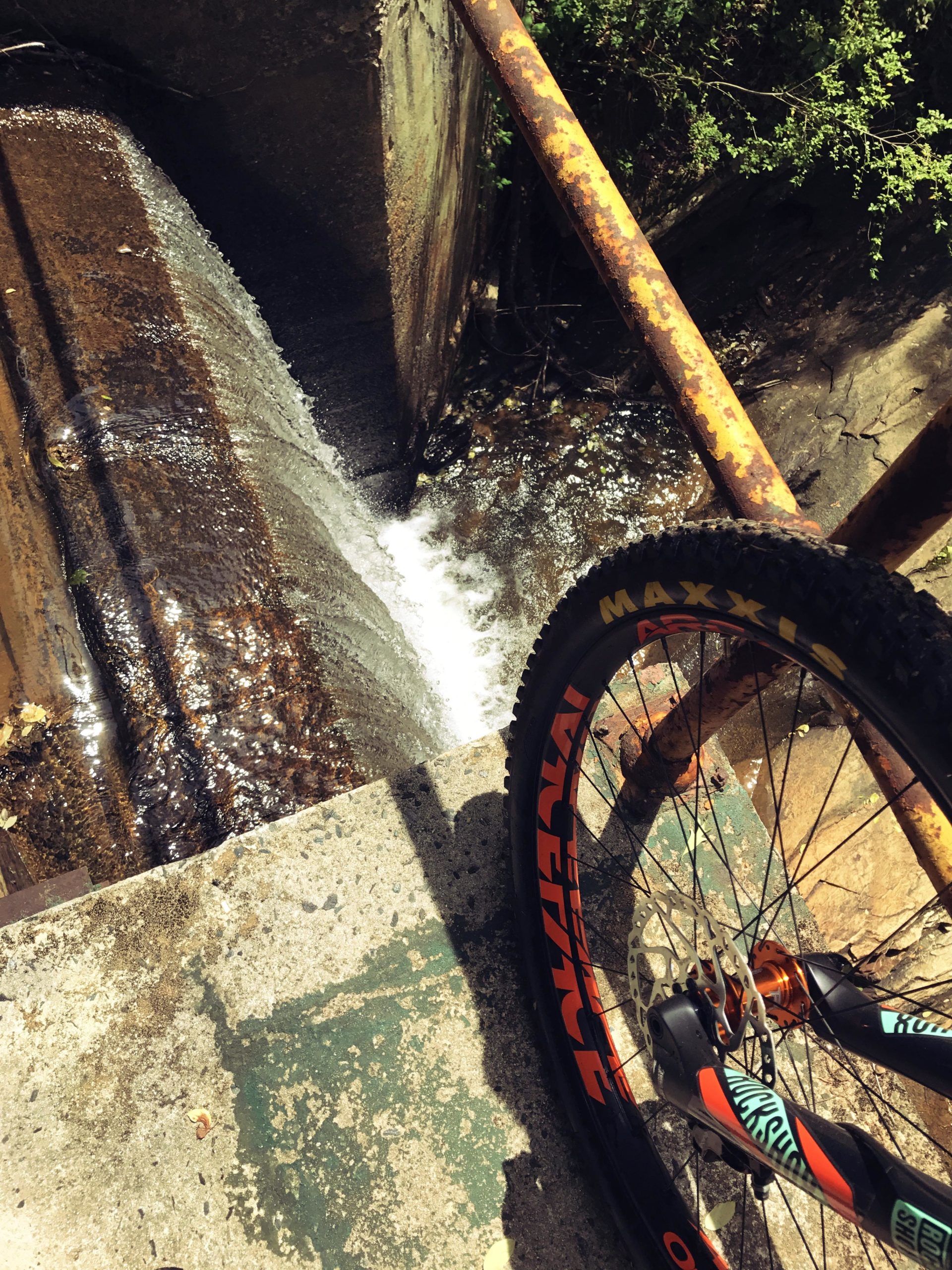 A close-up view of a mountain bike wheel resting on the edge of a concrete ledge above a small waterfall. The water cascades down into a narrow channel, surrounded by rocks and greenery. A rusty metal pipe runs along the top of the image, adding a contrast to the natural scene. Sunlight reflects off the water, creating a vibrant atmosphere. White Tail mountain bike trail.