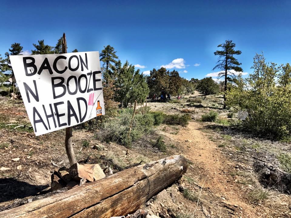 A wooden sign that reads "BACON N BOOZE AHEAD" is prominently displayed along a dirt path in a forested area. The background features trees under a blue sky with a few clouds, and some people can be seen in the distance along the path. The scene conveys a lighthearted and festive atmosphere. Sierra Prieta (Trail 366) mountain bike trail.