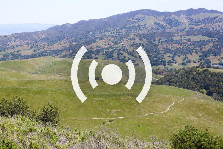 A wide view of rolling green hills and distant mountains under a clear blue sky, with a winding path visible through the landscape. An overlay symbol representing sound or audio is centered in the image.