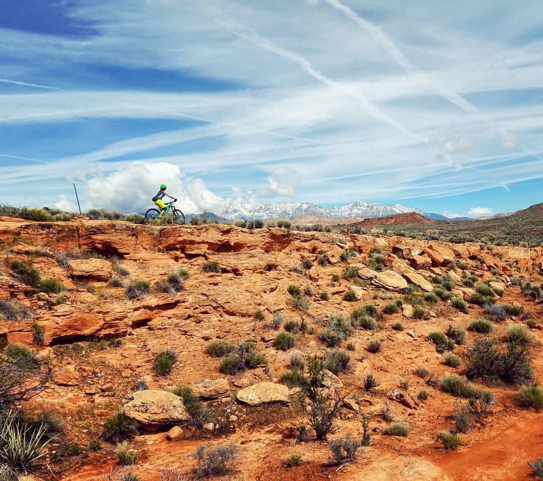 A mountain biker riding along a rocky trail in a desert landscape, with distant snow-capped mountains and a blue sky streaked with clouds in the background. The terrain features reddish-brown rocks and sparse vegetation, typical of a rugged outdoor environment. Prospector - Church Rocks Loop mountain bike trail.