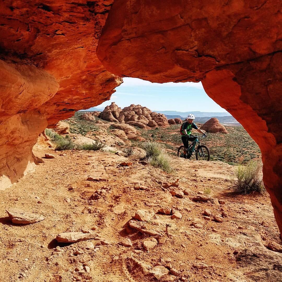 A cyclist wearing a helmet rides a mountain bike along a rocky trail framed by a natural arch of red rock. In the background, distinctive red rock formations rise against a blue sky, showcasing a rugged desert landscape. Prospector - Church Rocks Loop mountain bike trail.