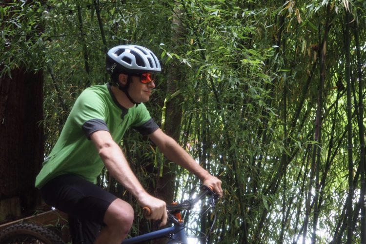 A person wearing a green shirt and a helmet, riding a mountain bike in a lush area surrounded by bamboo plants. The rider is looking ahead, suggesting focus and enjoyment of their outdoor activity.