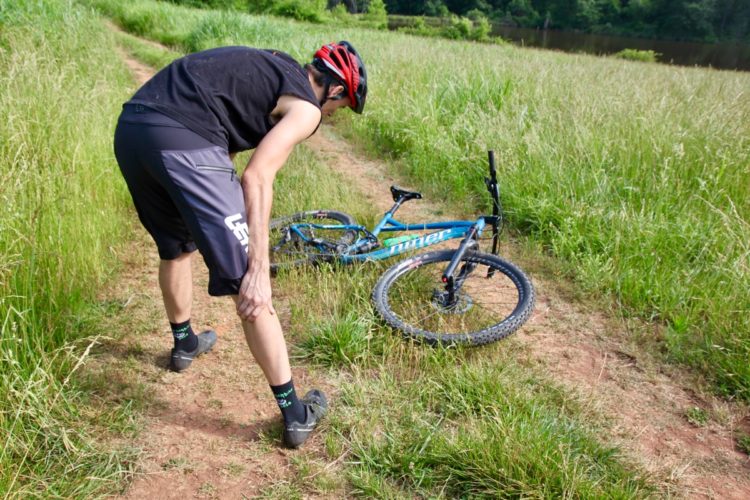 A person in athletic clothing is bending over on a dirt path, holding their leg, while a blue mountain bike lies on the ground nearby. The scene is surrounded by tall grass and trees, with a body of water visible in the background.