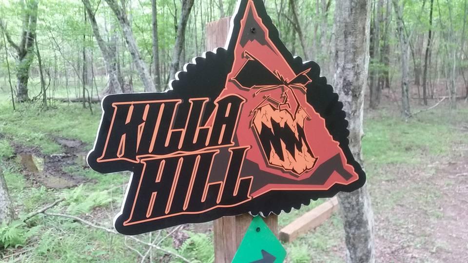 A triangular sign attached to a wooden post, featuring the text "KILLA HILL" in bold, stylized lettering. The sign includes a graphic of a fierce-looking creature or animal with an aggressive expression. The background features a lush, green forest setting with trees and foliage. Mountain Laurel Trails mountain bike trail.