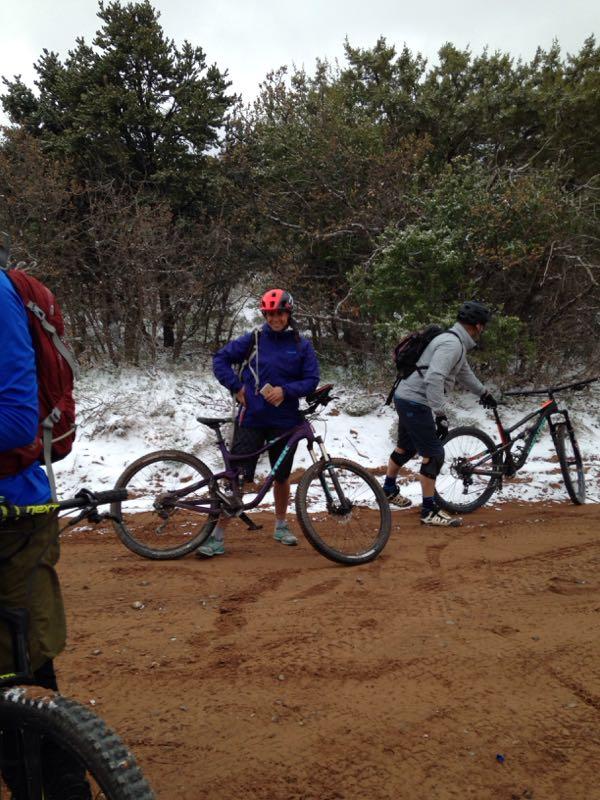 A group of mountain bikers stopped on a dirt trail, with snow covering the ground in some areas. One rider, wearing a purple jacket and a red helmet, stands beside a purple bicycle, while another rider in a gray sweatshirt and shorts walks with their bike in the background. The scene is surrounded by green shrubs and trees, indicating a wintery outdoor setting. Porcupine Rim mountain bike trail.