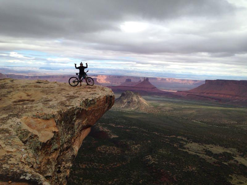 A mountain biker stands triumphantly on the edge of a rocky cliff, raising arms in celebration. The expansive desert landscape stretches out below, featuring mesas and canyons under a cloudy sky. Porcupine Rim mountain bike trail.