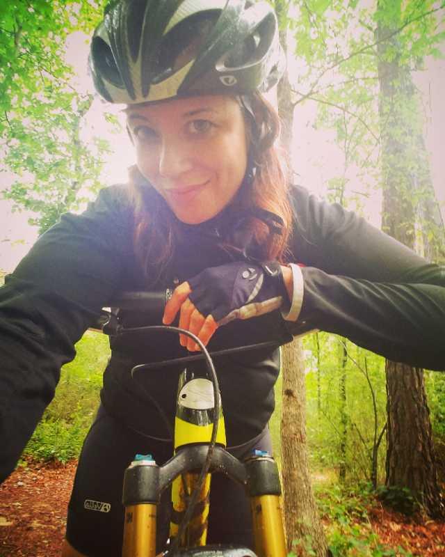 A person wearing a bicycle helmet and gloves smiles while posing for a selfie on a mountain bike. The background features lush green trees, indicating an outdoor trail setting. Trail Creek Park mountain bike trail.