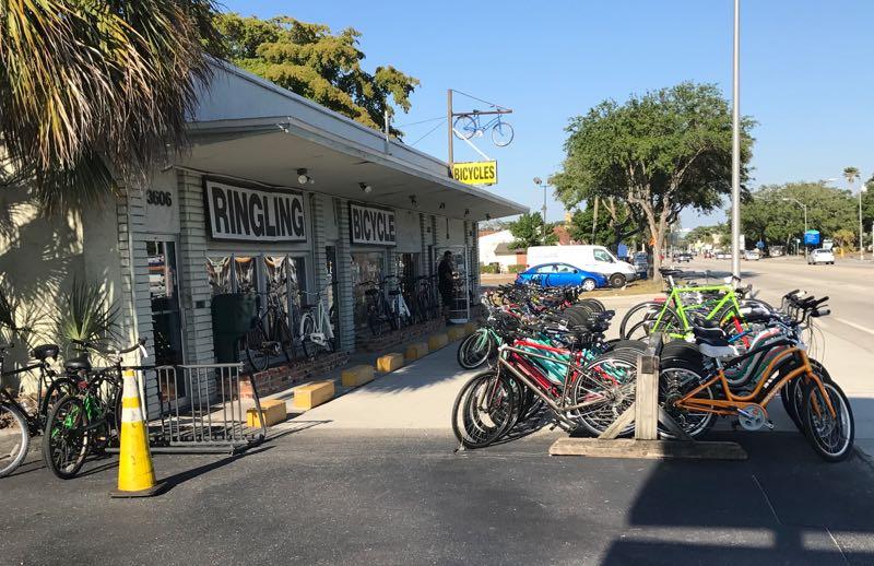 A bicycle shop named Ringling Bicycle with a variety of bicycles displayed outside. The building has large signage indicating the shop name, and several bicycles are parked along the side. The street is visible in the background, featuring greenery and a clear blue sky.