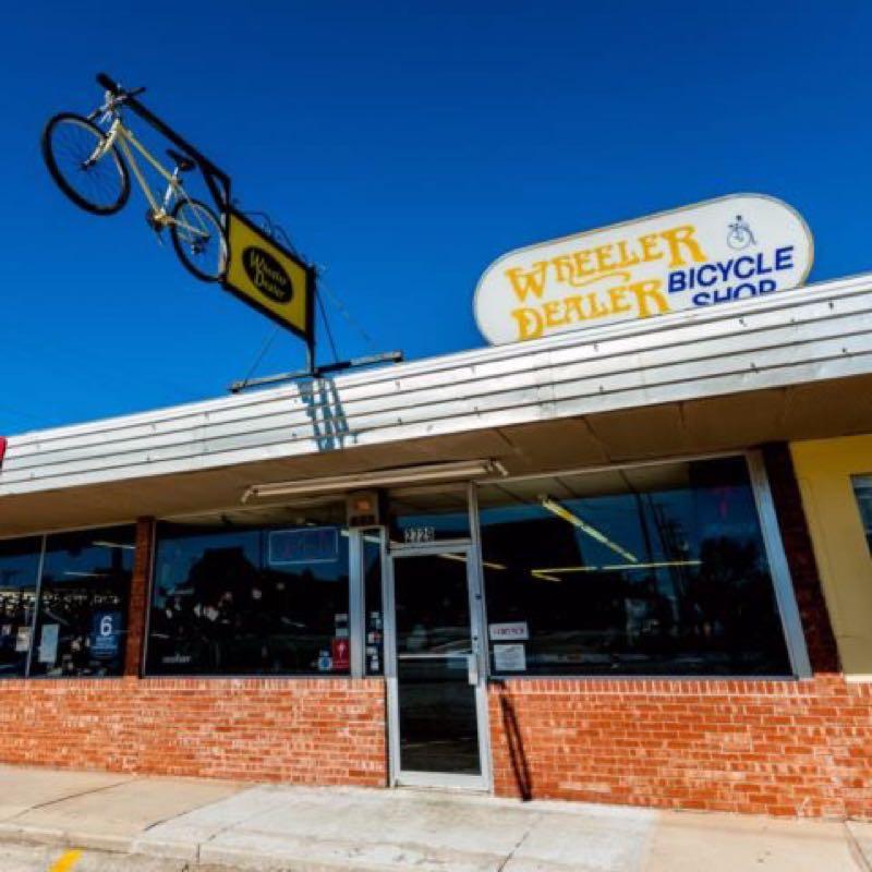 Storefront of the Wheeler Dealer Bicycle Shop, featuring a yellow bicycle mounted on a sign above the entrance and large glass windows. The brick façade contrasts with the white and yellow signage. Clear blue sky in the background.