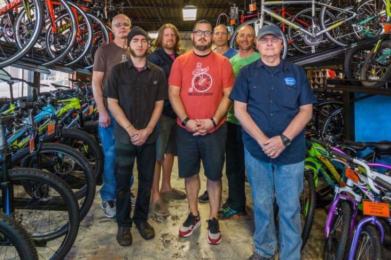 Group of seven individuals standing together in a bicycle shop, surrounded by an array of bicycles. The people are dressed casually and are positioned in two rows, with various bicycles hanging in the background. The setting appears to be a bike sales or repair shop, showcasing both mountain and road bikes.