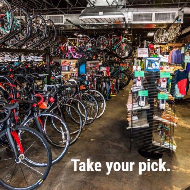 A brightly lit bike shop interior filled with various bicycles displayed on racks and the floor. Accessories and cycling apparel are visible on shelves and displays. The text "Take your pick." is prominently featured in the lower right corner.