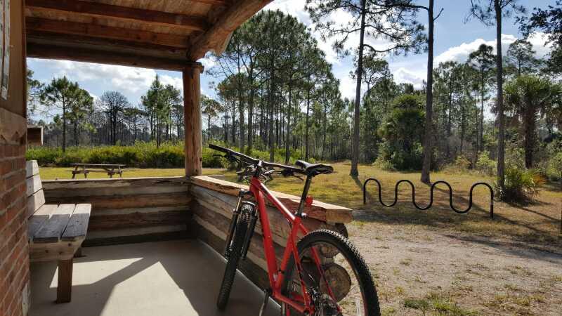 A red mountain bike rests against a wooden handrail on the porch of a rustic cabin. In the background, a grassy area is visible with picnic tables and a bike rack, surrounded by tall pine trees under a clear blue sky. Carlton Preserve mountain bike trail.