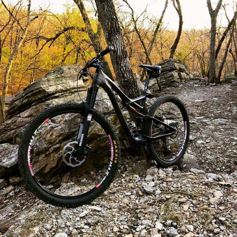A black mountain bike leaning against a tree near large rocks, surrounded by a forest with autumn foliage and a gravel path. The scene captures the natural landscape during golden hour. Swope Park Trail mountain bike trail.