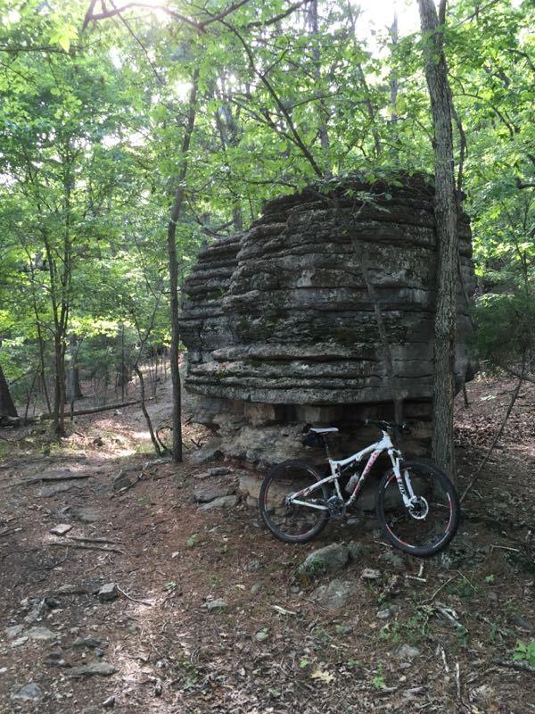 A mountain bike leaned against a large, round stone structure in a wooded area, with trees and underbrush surrounding the scene. Sunlight filters through the leaf canopy above, creating a serene outdoor atmosphere. Dam Overlook mountain bike trail.