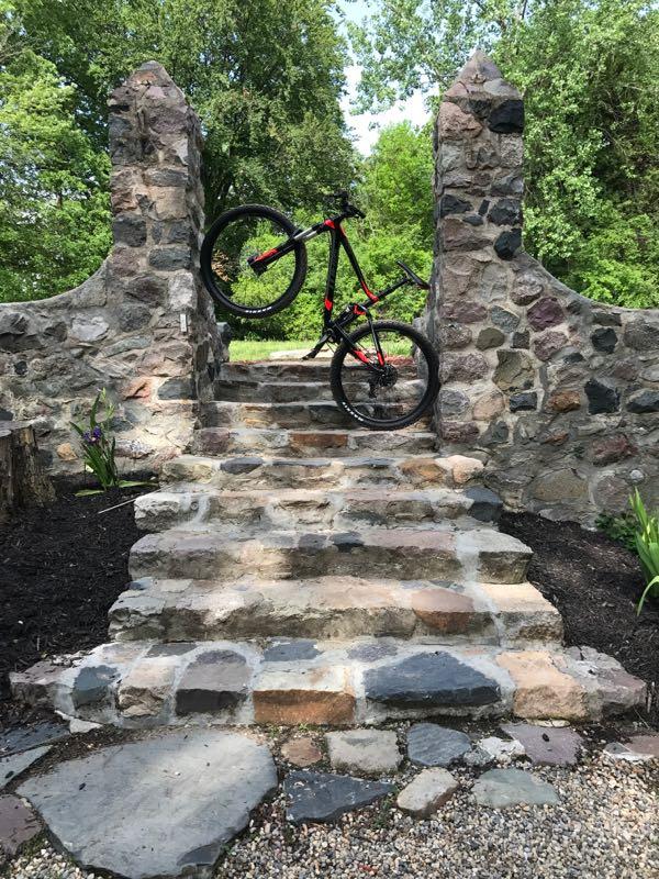 A mountain bike is positioned upright on a stone staircase leading through an archway made of weathered stones, with lush greenery in the background. The bike has black and red colors, and the staircase is flanked by decorative plants. Stony Creek Metro Park mountain bike trail.