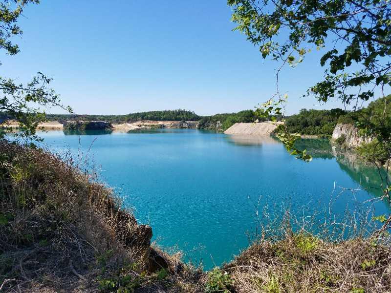 A serene view of a bright turquoise lake surrounded by green foliage and rocky cliffs under a clear blue sky. The water's calm surface reflects the vibrant colors of the landscape, creating a peaceful natural setting. Haile's Trails mountain bike trail.