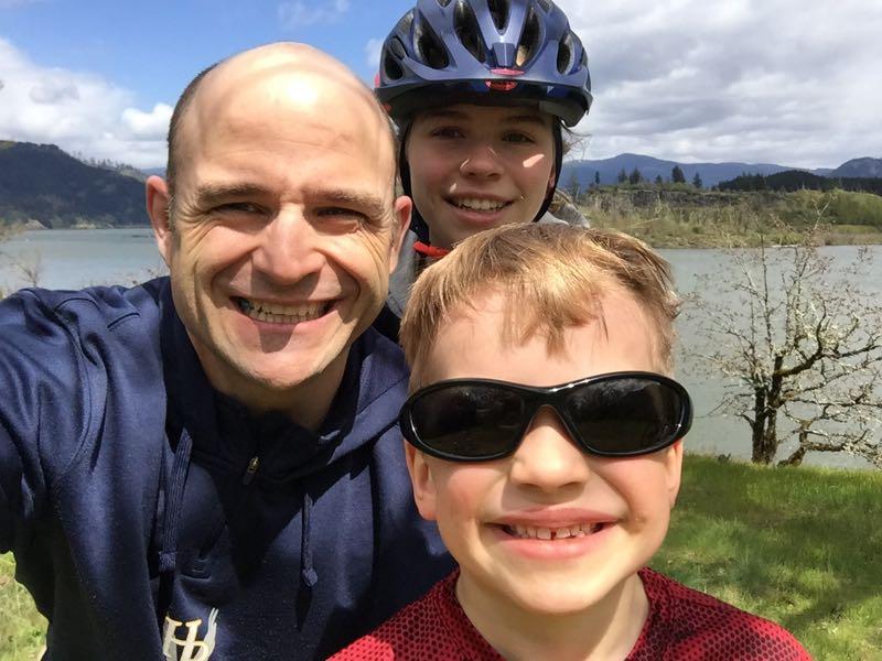 A smiling adult man and two children pose for a selfie outdoors by a lake. The man is wearing a dark hoodie, while one child has on sunglasses and the other is wearing a bike helmet. The background features green hills and a partly cloudy sky. easyCLIMB mountain bike trail.