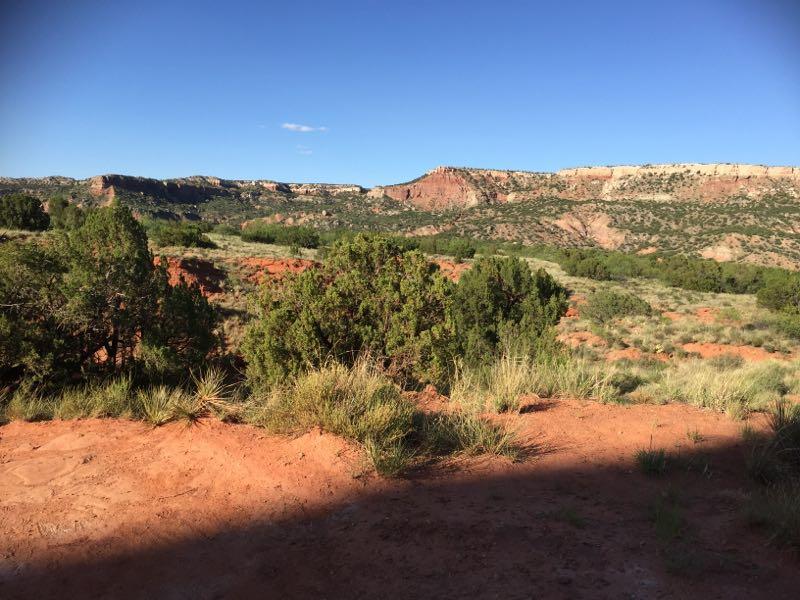 A scenic view of a rugged landscape featuring red rock formations and green shrubs under a bright blue sky. The terrain includes rolling hills and distant cliffs, showcasing the natural beauty of the area. Palo Duro Canyon mountain bike trail.