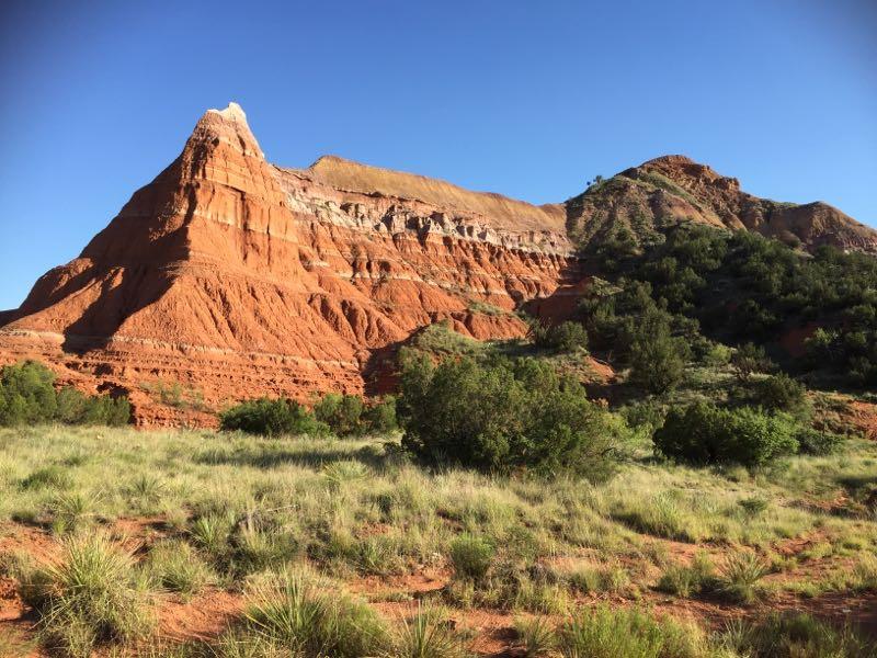 A striking red rock formation rises against a clear blue sky, surrounded by green vegetation and grassland in the foreground. The layers of the rock display various shades and textures, indicating geological history. Palo Duro Canyon mountain bike trail.