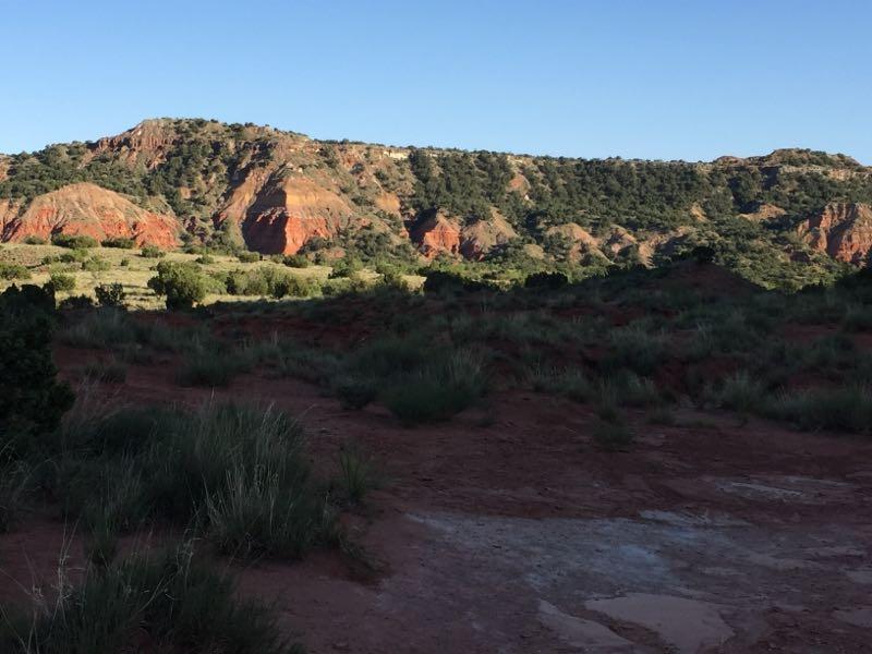 A scenic view of red rock formations and hills under a clear blue sky, surrounded by greenery and open land. The landscape captures the natural beauty of the terrain, showcasing varying shades of red and green. Palo Duro Canyon mountain bike trail.