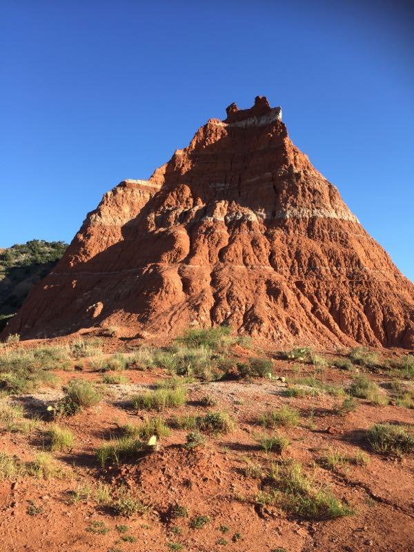 A steep, reddish-brown rock formation stands prominently against a clear blue sky, showcasing layered textures and contours. Sparse green vegetation grows at the base, accentuating the natural landscape. Palo Duro Canyon mountain bike trail.