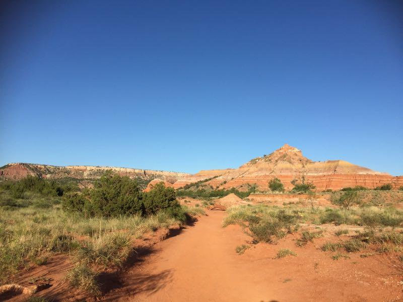 A dirt path meanders through a grassy area leading towards a prominent rocky hill under a clear blue sky. The landscape features shades of red and orange cliffs in the background, surrounded by sparse greenery and small bushes. Palo Duro Canyon mountain bike trail.