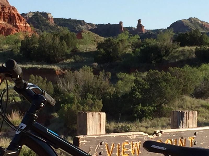 A scenic view of a rugged landscape featuring red rock formations and green foliage, with a bike in the foreground next to a wooden sign that reads "VIEW POINT." The sunlight casts a warm glow on the scenery, showcasing the natural beauty of the area. Palo Duro Canyon mountain bike trail.