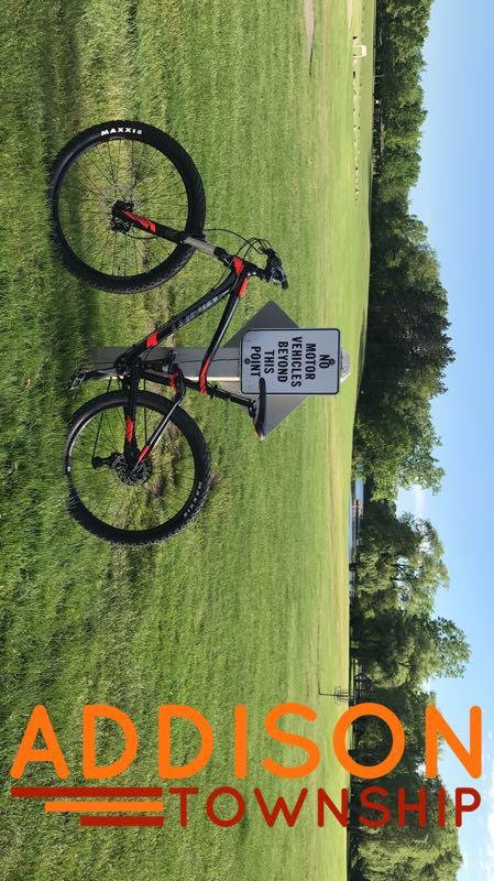 A mountain bike is leaning against a sign that reads "No motor vehicles beyond this point." The background features a grassy field and trees, indicating a park or outdoor recreational area. The text "ADDISON TOWNSHIP" is prominently displayed at the bottom of the image. Addison Oaks mountain bike trail.
