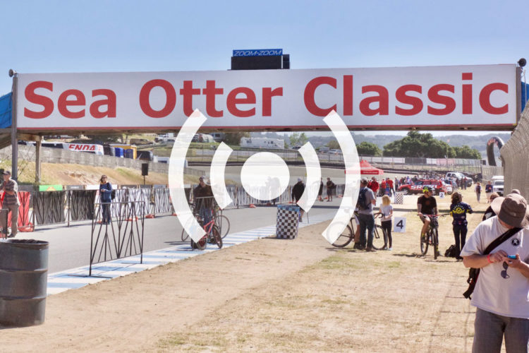 A wide view of the Sea Otter Classic event entrance, featuring a large banner with bold red lettering. In the foreground, people of various ages are seen near the race track, some on bicycles and others walking. Nearby, spectators and participants gather, with a backdrop of tents and vehicles in the distance, indicating a lively outdoor event atmosphere.