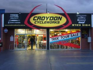 A storefront of Croydon Cycleworks, featuring large signage with the business name and logos of various bike brands. The entrance is illuminated, with a person walking in. A promotional banner for a Trek tour sale is prominently displayed in the window. The scene is set during twilight, highlighting the shop's exterior.