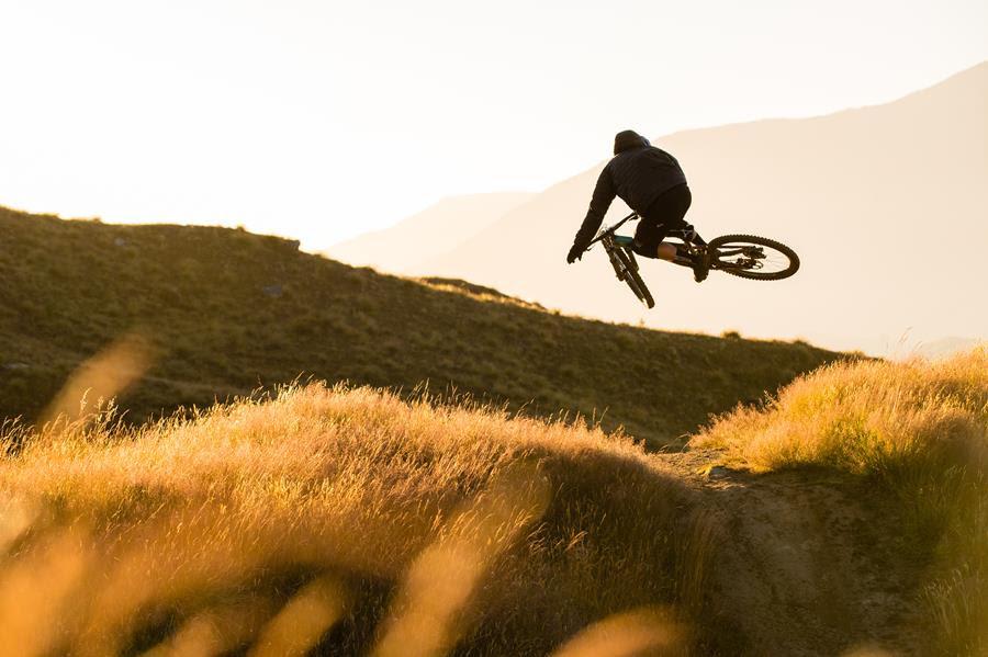 Rocky Mountain Altitude: A mountain biker performing a jump over a dirt mound in a golden sunset landscape, with grassy hills and distant mountains in the background.