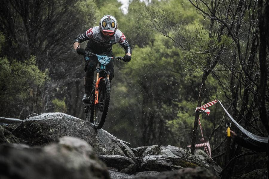 Rocky Mountain Altitude: A mountain biker wearing a colorful helmet and jersey rides over rocky terrain while it's raining, surrounded by lush greenery. The biker is airborne, and a red and white caution tape can be seen in the background, indicating a course or path.