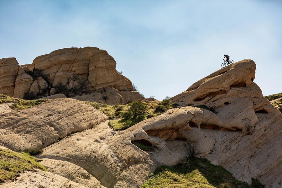 Rocky Mountain Altitude: A mountain biker riding on a rocky outcrop against a clear blue sky, surrounded by rugged terrain and green vegetation.