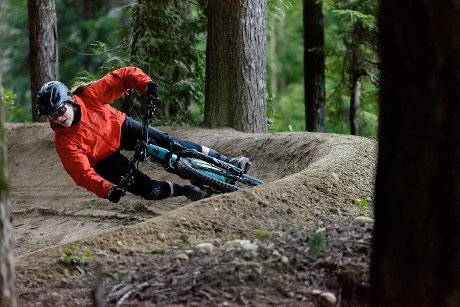 Rocky Mountain Altitude: A person in an orange jacket and helmet rides a mountain bike along a dirt track, leaning into a turn amidst a forest of tall trees. The scene captures the thrill of biking on a winding trail surrounded by nature.