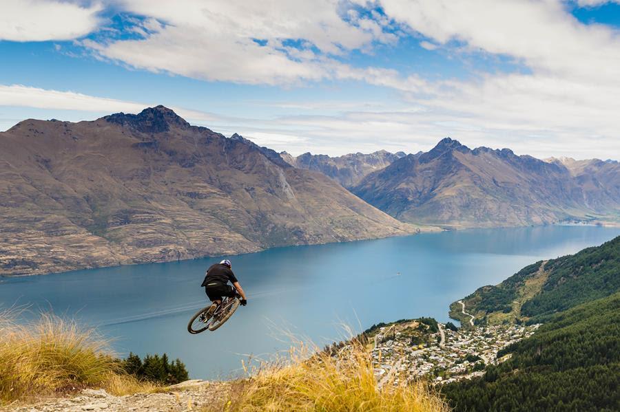 Rocky Mountain Altitude: A mountain biker performs a jump on a dirt trail overlooking a scenic lake and mountainous landscape under a partly cloudy sky. The vibrant blue water contrasts with the rugged terrain and lush greenery below, showcasing the beauty of outdoor adventure.